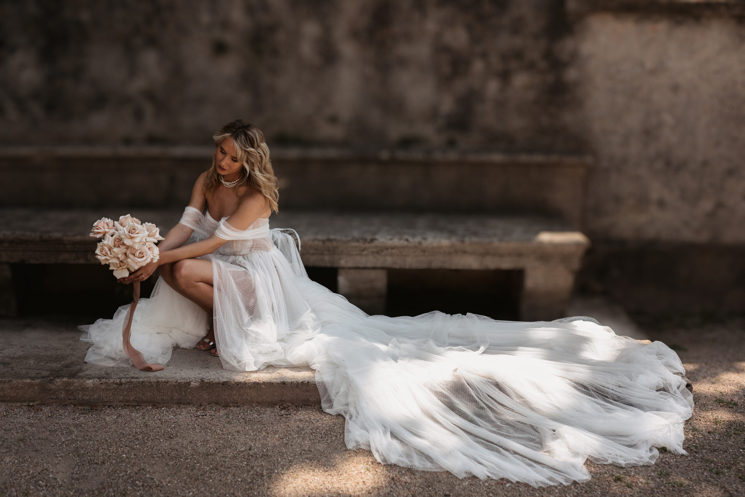 Braut mit Rosenstrauß und langer Schleppe auf einer Steinbank – Hochzeitsfotografie von Kenan Egin
