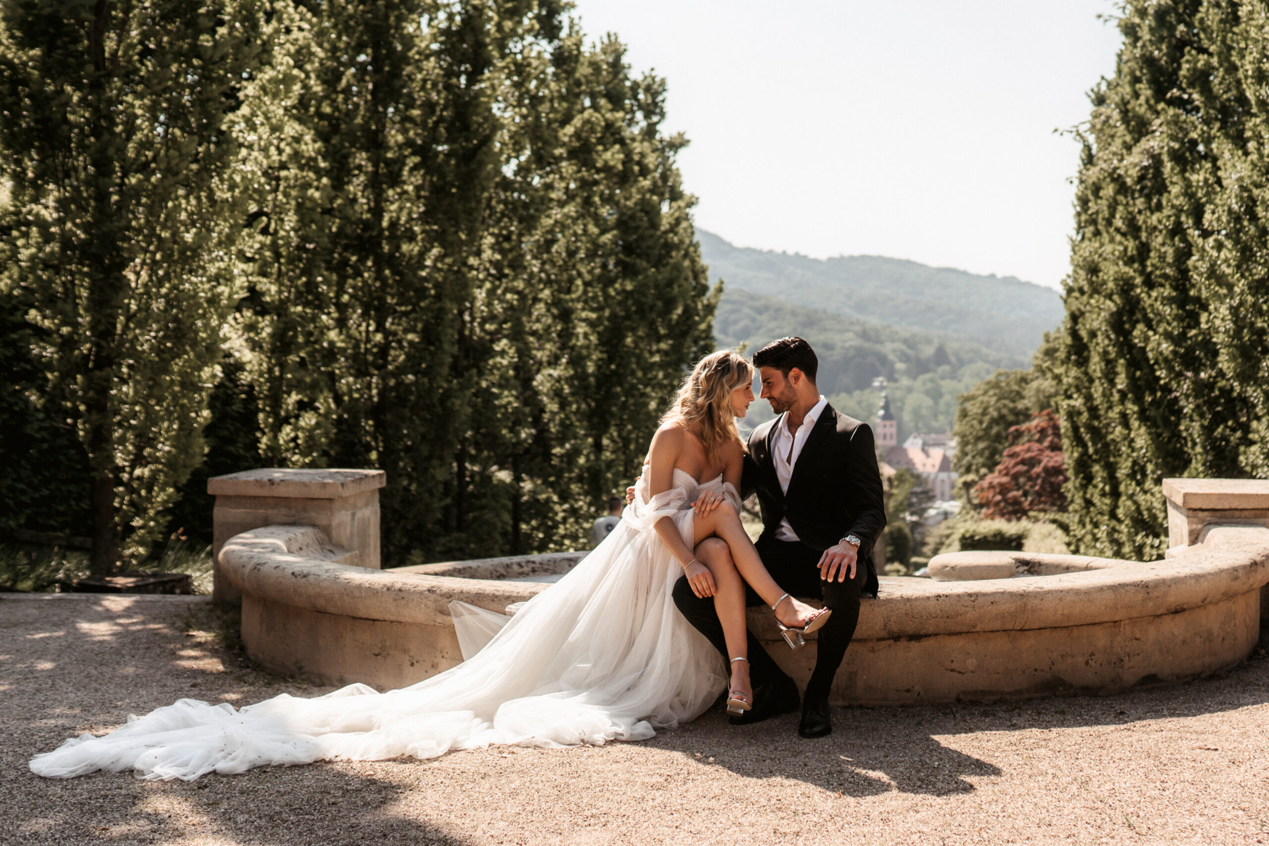 Braut und Bräutigam sitzen einander zugewandt zwischen hohen Zypressen – Hochzeitsfotografie von Kenan Egin