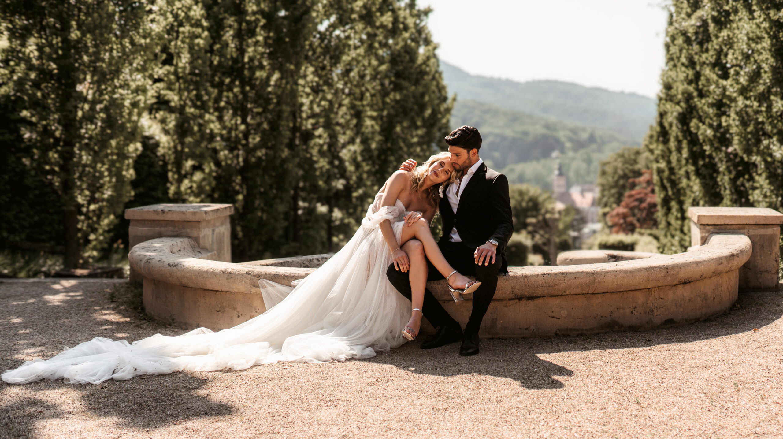 Braut lehnt ihren Kopf an den Bräutigam auf einer steinernen Gartenbank – Hochzeitsfotografie von Kenan Egin