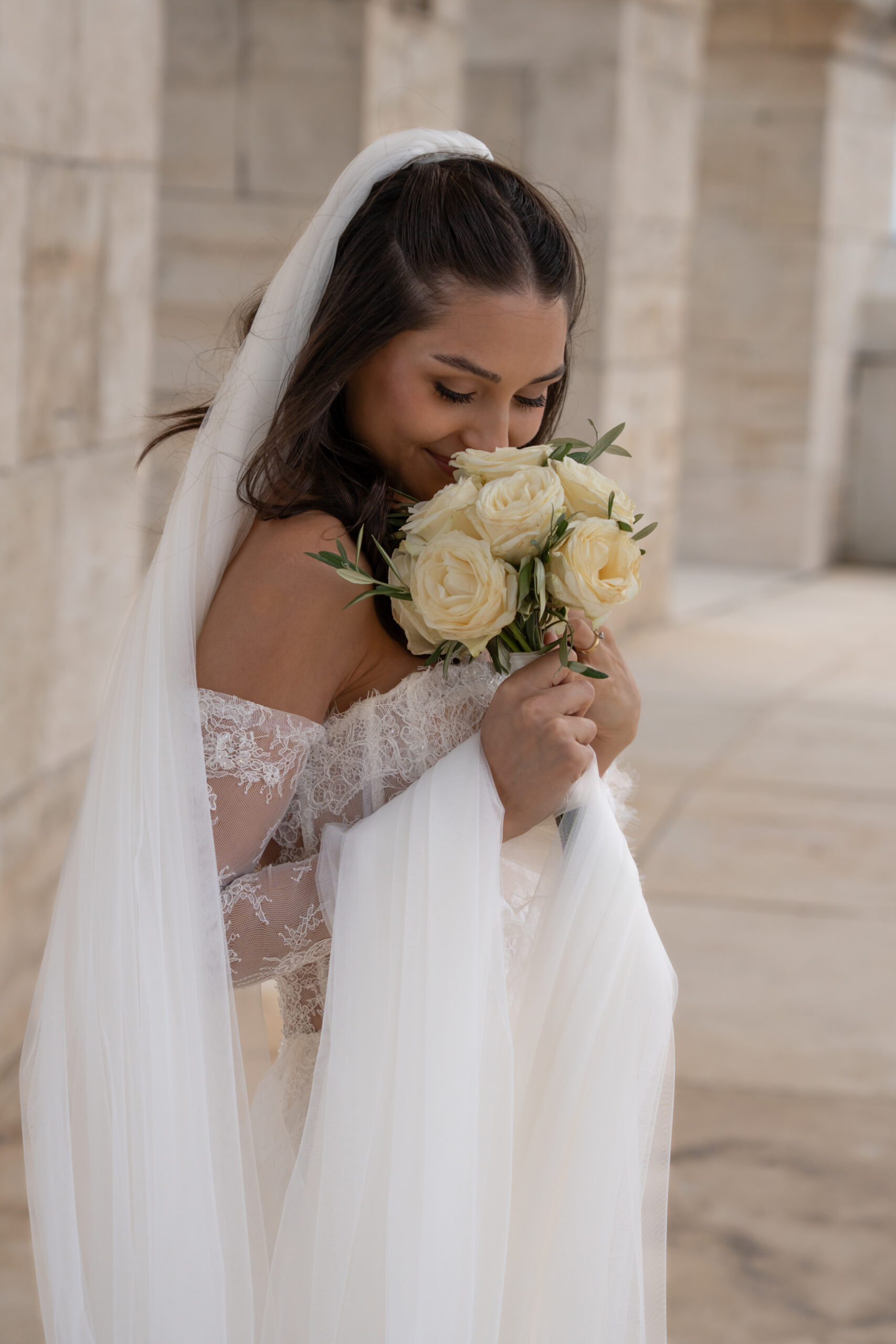 Braut im Spitzenkleid riecht an ihrem Strauß aus weißen Rosen.